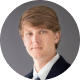 Headshot of a young man with side-swept light brown hair wearing a dark suit and tie, set against a neutral gray background. He has a serious, confident expression.