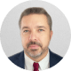 Headshot of a man in a suit and red tie with a short beard, set against a light gray background.