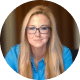 A woman with long blonde hair and glasses, wearing a blue shirt, sits indoors in front of a blurred background, looking directly at the camera with a slight smile.